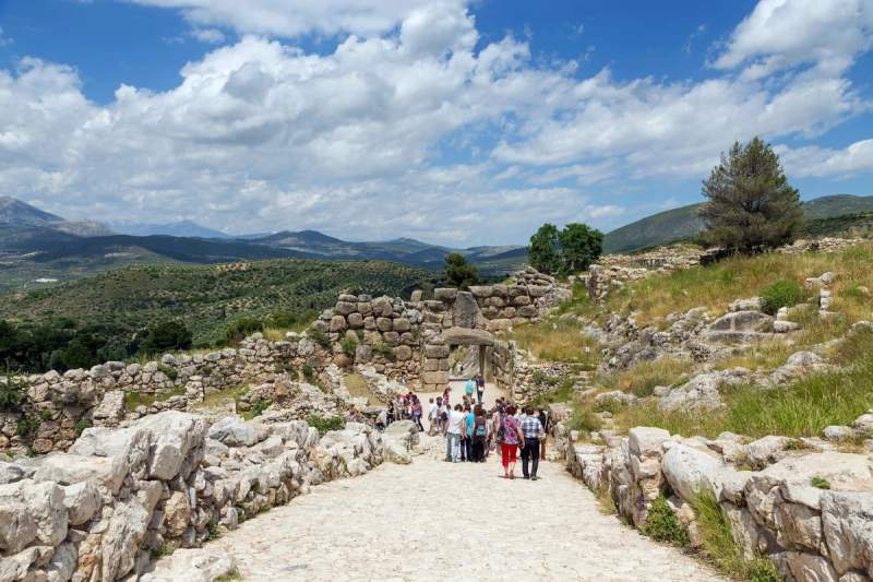 Vue du site archéologique de Mycènes avec des visiteurs marchant vers la Porte des Lionnes, entourée de vestiges en pierre et de collines verdoyantes sous un ciel bleu.
