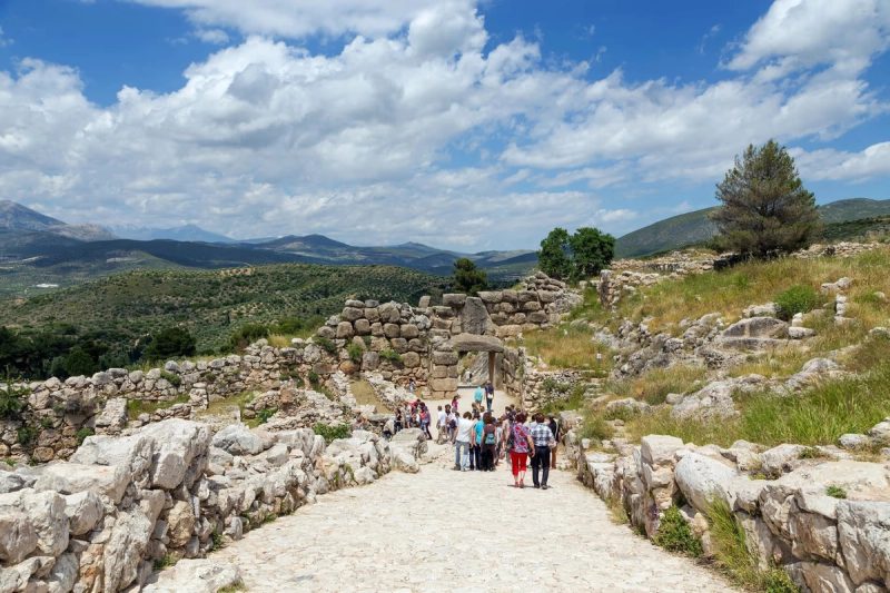 Vue du site archéologique de Mycènes avec des visiteurs marchant vers la Porte des Lionnes, entourée de vestiges en pierre et de collines verdoyantes sous un ciel bleu.