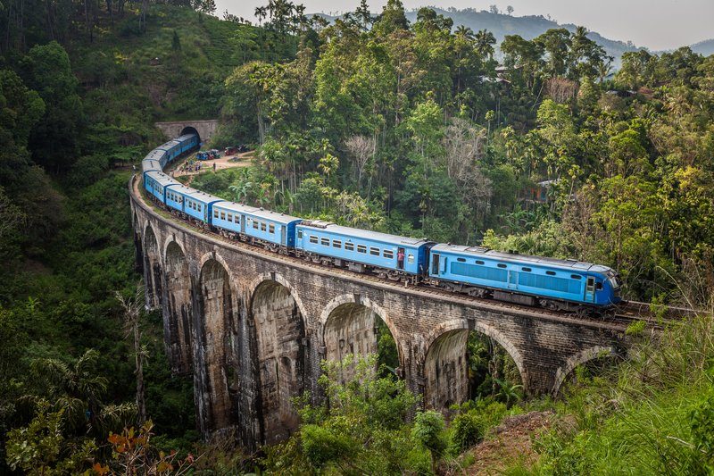 Pont ferroviaire en pierre à neuf arches traversé par un train bleu, entouré d’une jungle dense.