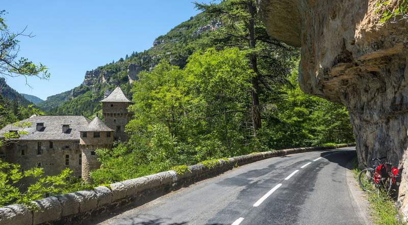 Route sinueuse bordée de falaises et forêt, avec un château en pierre niché dans les gorges des Cévennes.