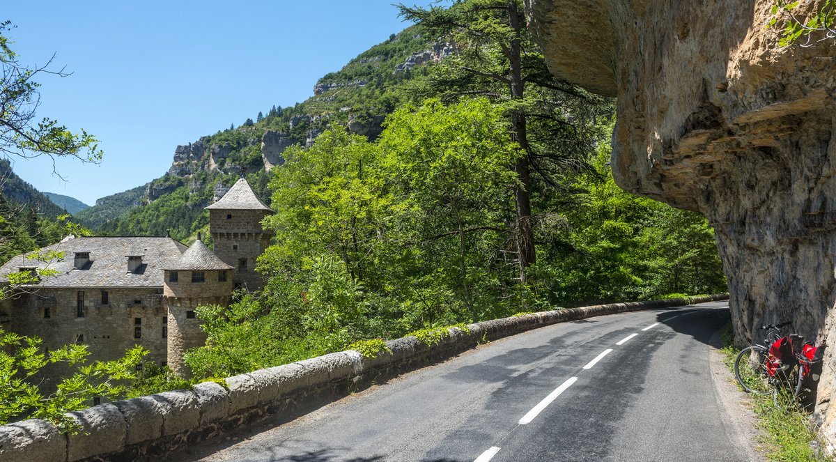 Route sinueuse bordée de falaises et forêt, avec un château en pierre niché dans les gorges des Cévennes.