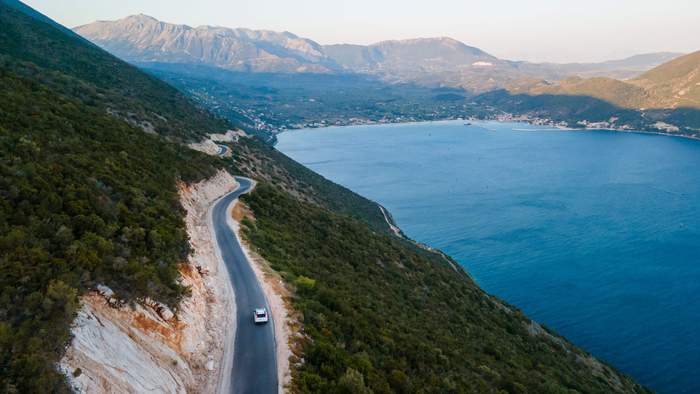 Vue aérienne d’une route côtière sinueuse en Épire, au nord-ouest de la Grèce, longeant une montagne verdoyante avec la mer Ionienne en contrebas, parfaite pour une micro-aventure estivale loin des foules.