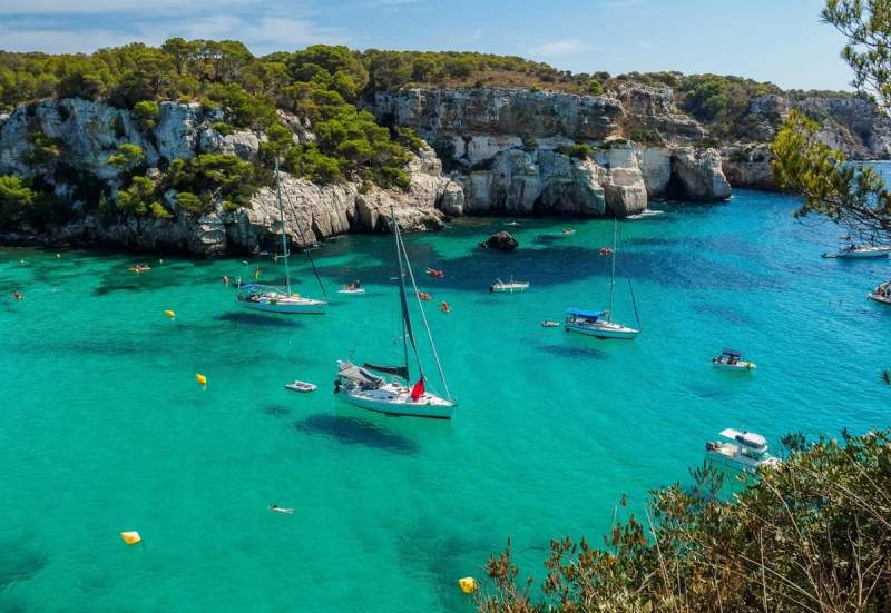 Vue panoramique de Cala Macarella à Minorque avec ses eaux cristallines et turquoises, entourée de falaises verdoyantes. Plusieurs voiliers et bateaux sont ancrés dans la baie, profitant d'une journée ensoleillée.