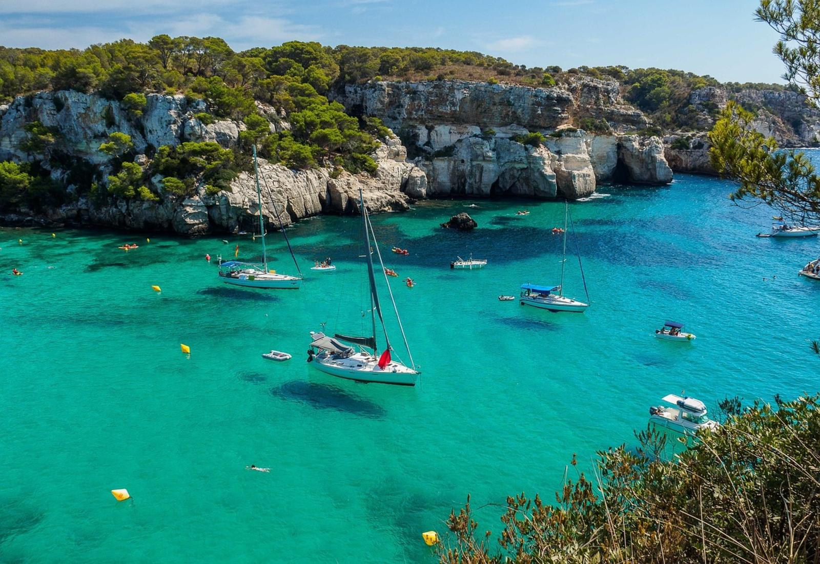 Vue panoramique de Cala Macarella à Minorque avec ses eaux cristallines et turquoises, entourée de falaises verdoyantes. Plusieurs voiliers et bateaux sont ancrés dans la baie, profitant d'une journée ensoleillée.