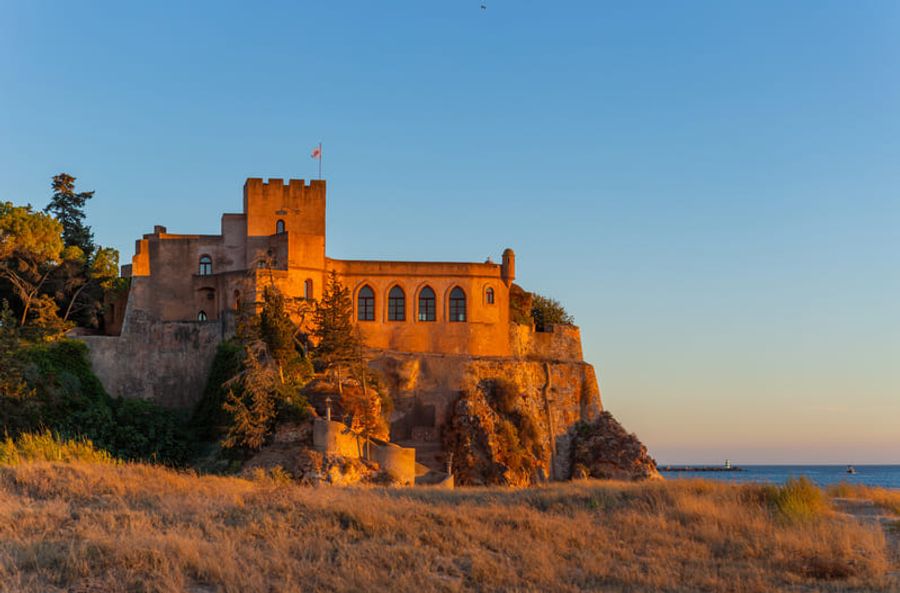 Un château médiéval en pierre ocre perché sur une falaise, avec une vue surplombant la mer, illuminé par le coucher de soleil.