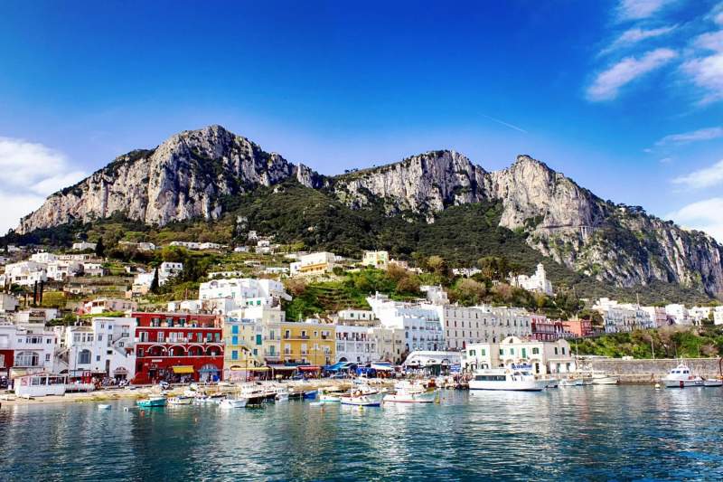 Vue sur les maisons colorées et le port de Capri avec les montagnes en arrière-plan, sous un ciel bleu éclatant.