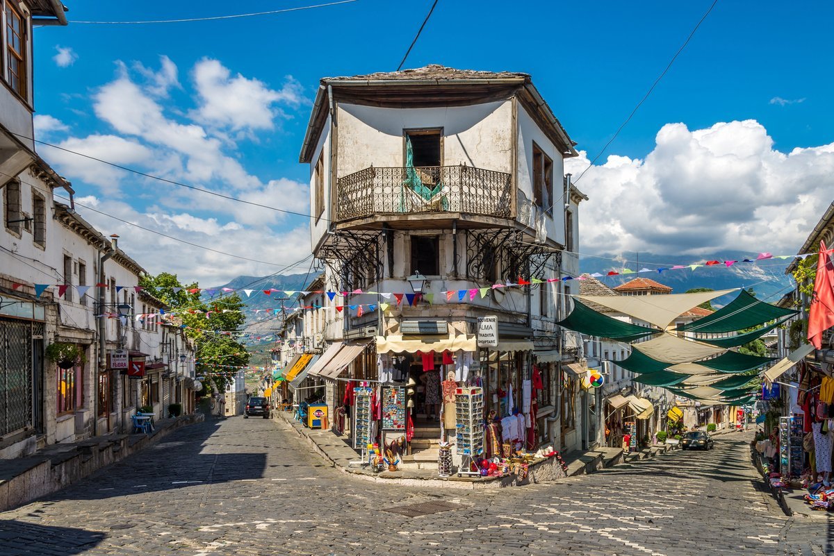 Rue pavée de la vieille ville de Gjirokastër en Albanie, bordée de maisons ottomanes traditionnelles et d'une boutique de souvenirs, avec les montagnes en arrière-plan