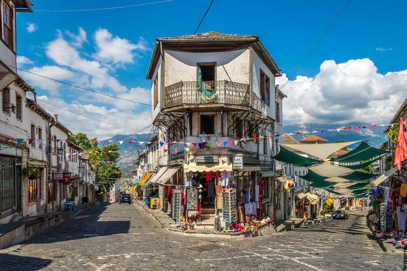 Rue pavée de la vieille ville de Gjirokastër en Albanie, bordée de maisons ottomanes traditionnelles et d'une boutique de souvenirs, avec les montagnes en arrière-plan