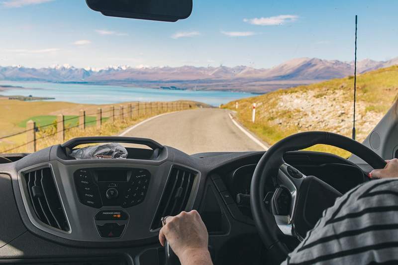 Vue depuis un van sur une route de Nouvelle-Zélande longeant un lac turquoise et des montagnes, lors d’un road trip en camping-car.