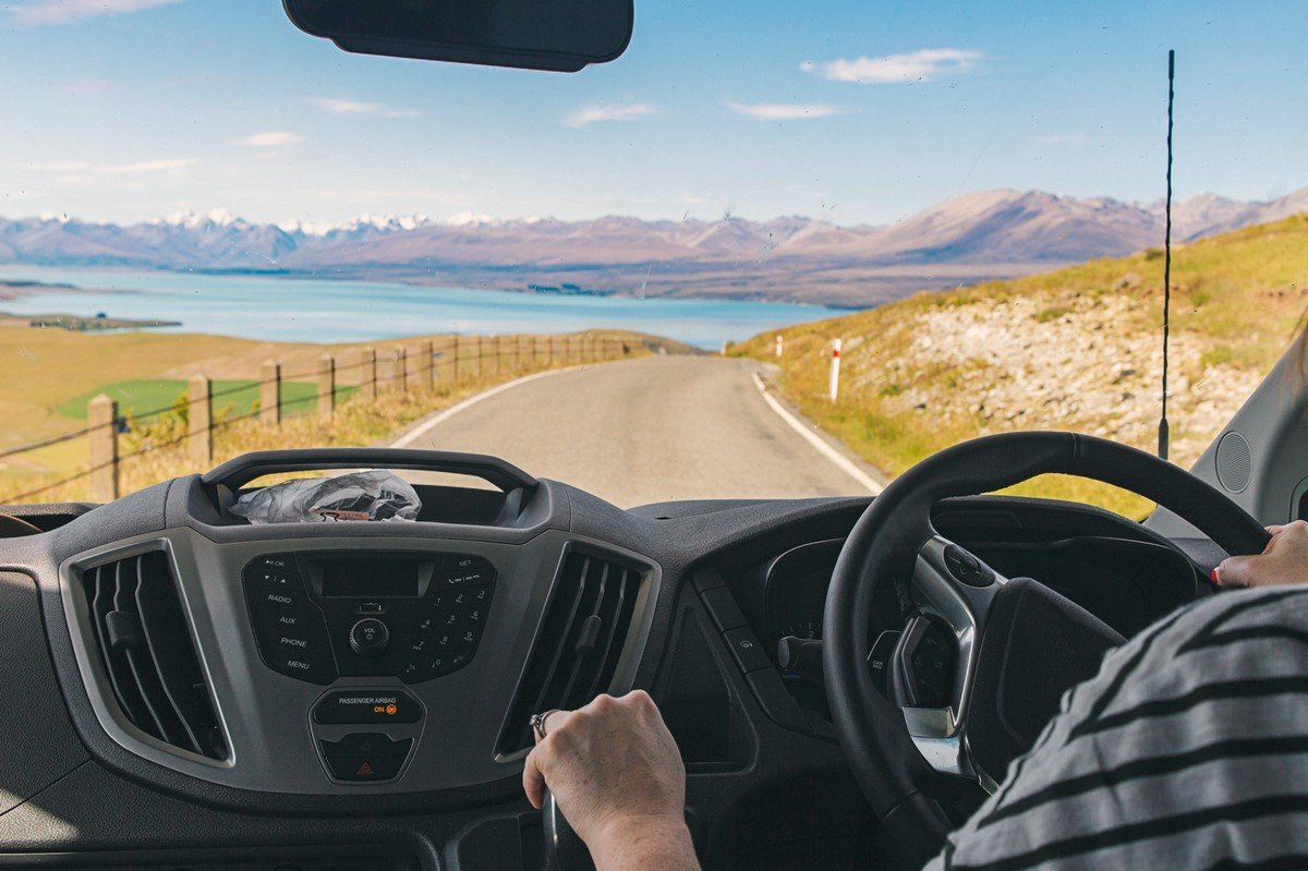 Vue depuis un van sur une route de Nouvelle-Zélande longeant un lac turquoise et des montagnes, lors d’un road trip en camping-car.