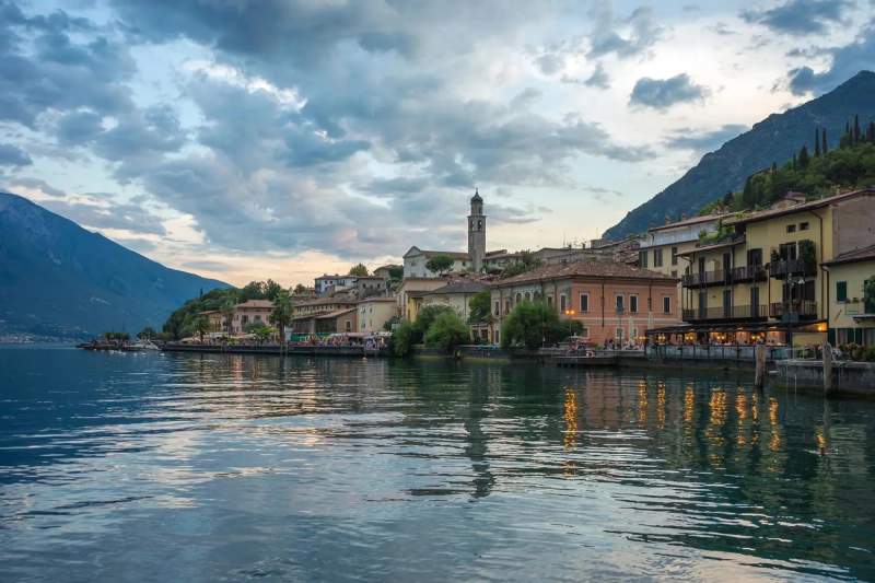 Vue panoramique d'un port au lac de garde en Italie avec des bâtiments colorés au bord de l'eau et des montagnes en arrière-plan.