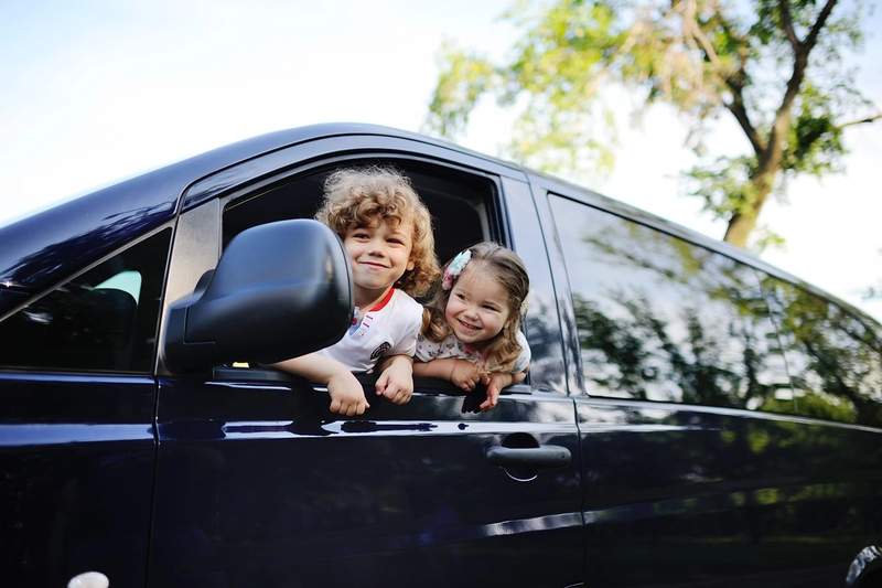 Deux jeunes enfants souriants regardent par la fenêtre d’un van aménagé lors d’un road trip en pleine nature.