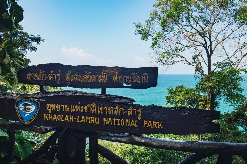 Panneau en bois indiquant l’entrée du Parc National de Khao Lak-Lam Ru avec vue sur l’océan turquoise.