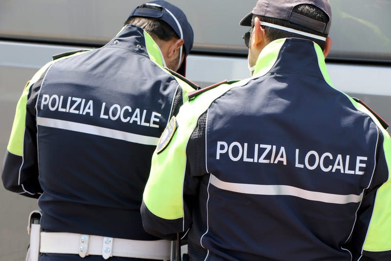 Deux agents de la police locale italienne vus de dos, en uniforme bleu et jaune fluorescent, en service dans la rue.