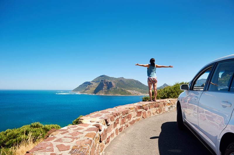 Voyageuse les bras écartés devant sa voiture de location garée sur une route côtière en bord de mer