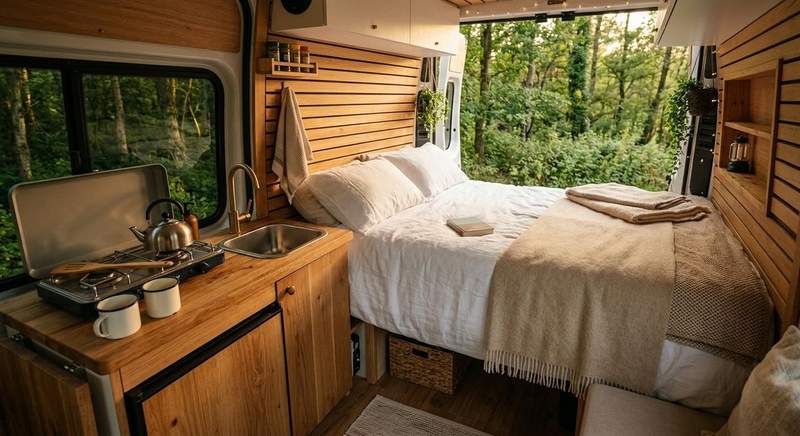 Intérieur aménagé d'un van avec lit double, cuisine en bois, réchaud et évier, vue sur la forêt