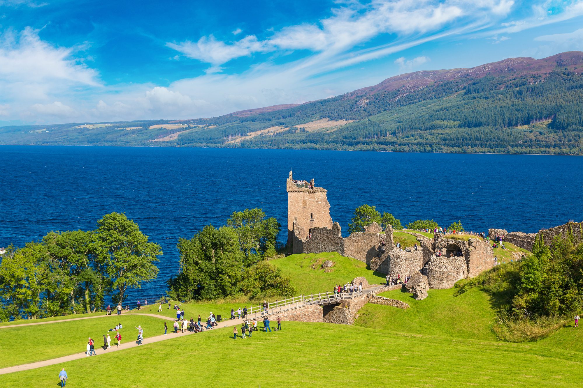 Les ruines et la tour d’Urquhart Castle dominent le Loch Ness, avec des visiteurs sur les chemins et les collines boisées sur la rive opposée