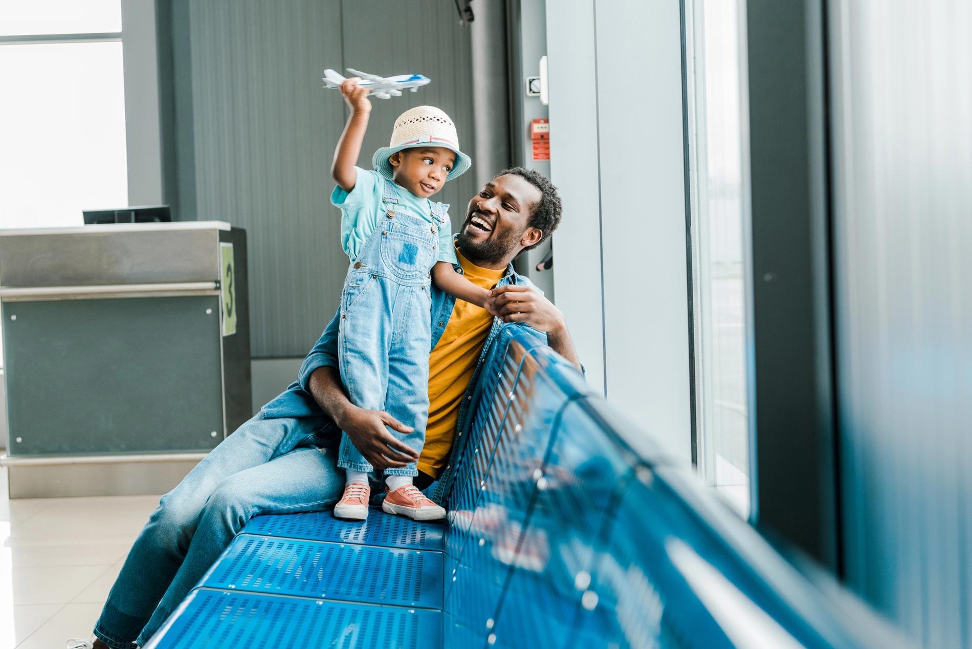 Un père et son enfant sourient ensemble dans un aéroport, l'enfant jouant avec un avion jouet près d'une baie vitrée.