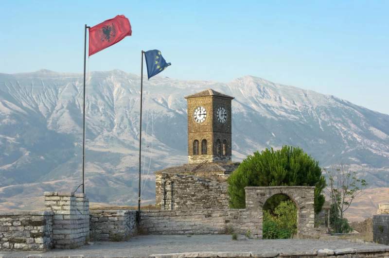 Tour de l’horloge historique en Albanie avec des drapeaux flottant au vent, encadrée par des montagnes majestueuses sous un ciel dégagé.