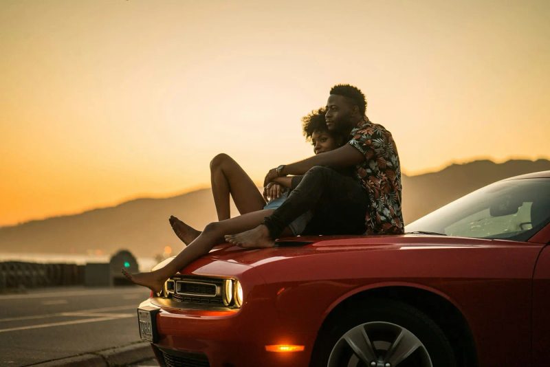 Un homme et une femme assis sur le capot d'une une voiture rouge au coucher du soleil