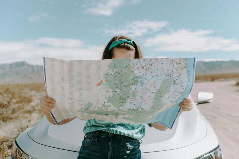 Une voyageuse se tenant debout devant une voiture tient une carte ouverte, à la recherche de son prochain arrêt sur un road trip.
