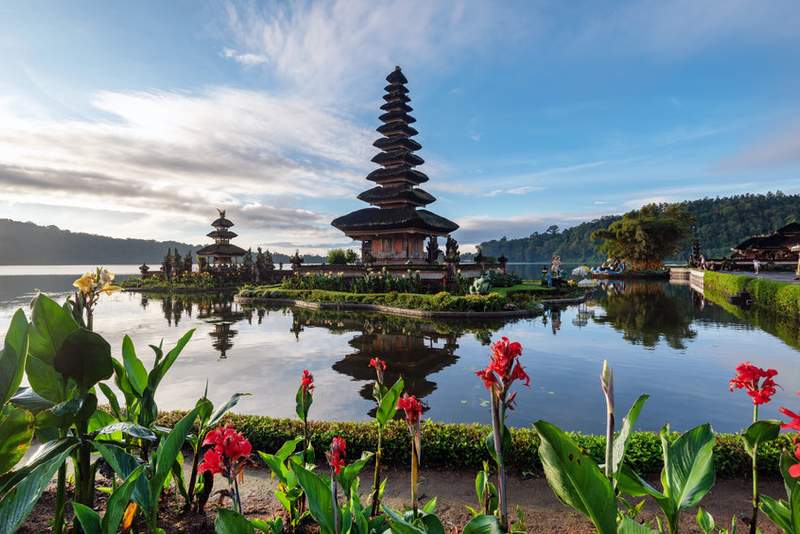 Le temple Ulun Danu Bratan à Bali, situé au bord du lac Bratan, entouré d'eau calme, de montagnes verdoyantes et de fleurs rouges.