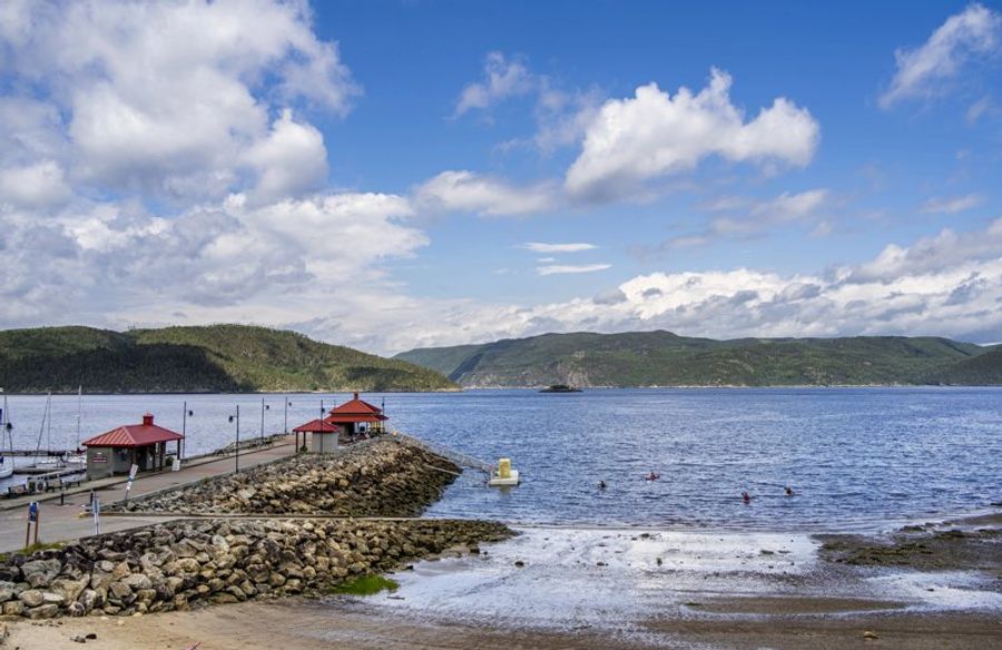 Vue panoramique du village de Tadoussac avec sa baie et ses bâtiments au toit rouge, entourés de collines verdoyantes.