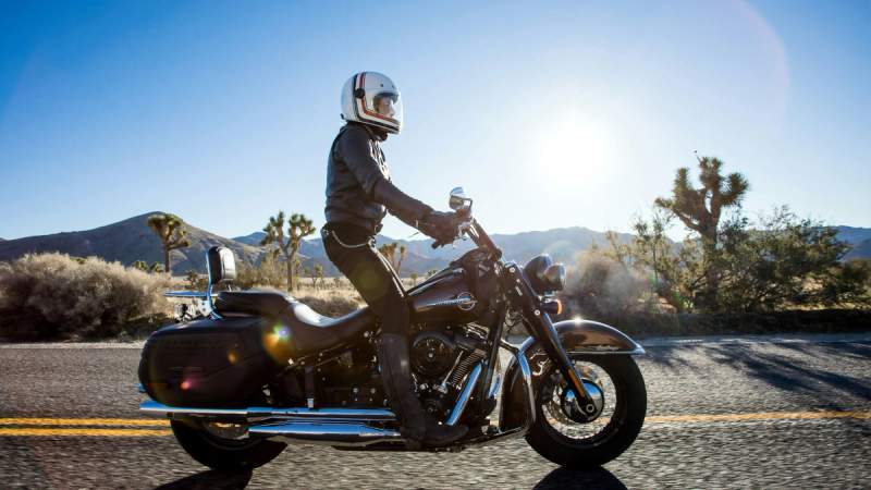 Un motard roulant sur une route désertique avec une Harley-Davidson, vêtu d'une veste en cuir noir et portant un casque blanc. Le paysage environnant est composé d'arbustes, de quelques arbres, et de montagnes au loin, sous un ciel bleu clair.