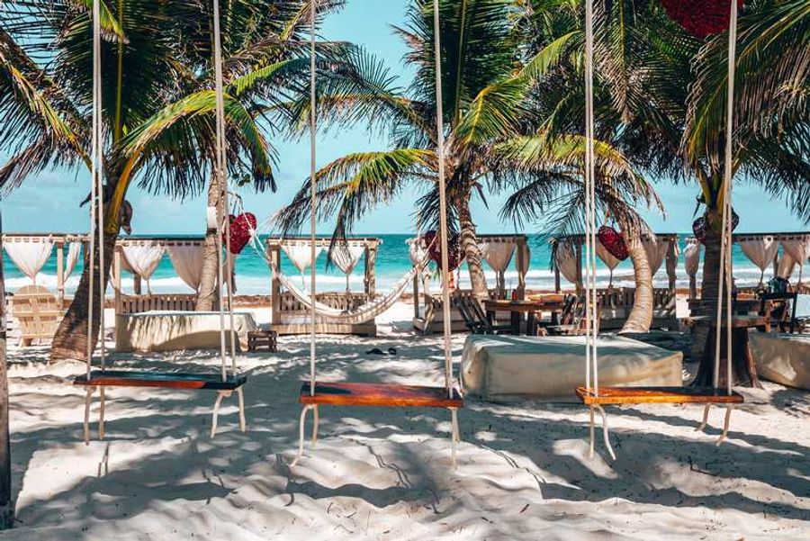 Plage de sable blanc à Tulum avec balançoires, palmiers, lits balinais et cabanes en toile face à la mer turquoise.