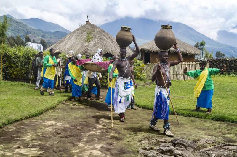 Scène de village traditionnel rwandais avec procession en habits colorés devant des huttes en chaume, sur fond de montagnes verdoyantes.