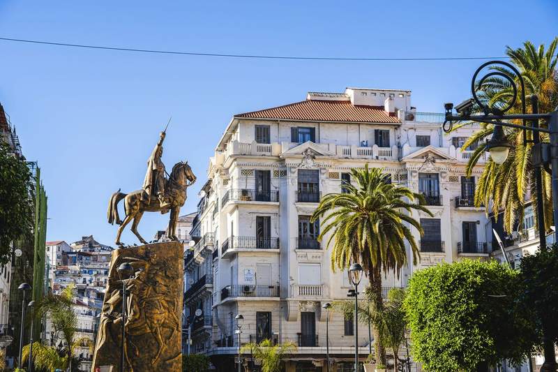 Statue de l’émir Abdelkader à cheval au centre-ville d’Alger, entourée d’immeubles haussmanniens et de palmiers sous un ciel bleu clair.