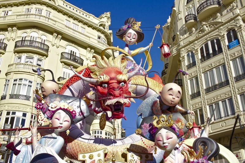Sculptures colorées et géantes exposées lors de la fête des Fallas à Valence, représentant un dragon rouge et des personnages asiatiques dans une rue animée.