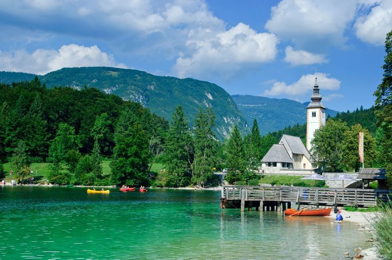 Lac de Bohinj en Slovénie avec ses eaux turquoise, des kayaks colorés et l'église Saint-Jean-Baptiste en arrière-plan