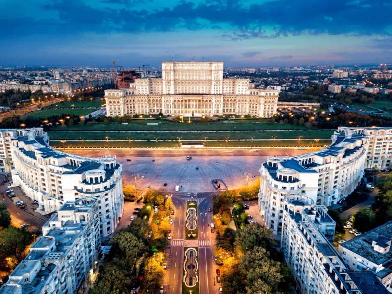 Vue aérienne du Palais du Parlement à Bucarest au crépuscule, entouré de bâtiments et d'avenues illuminées.