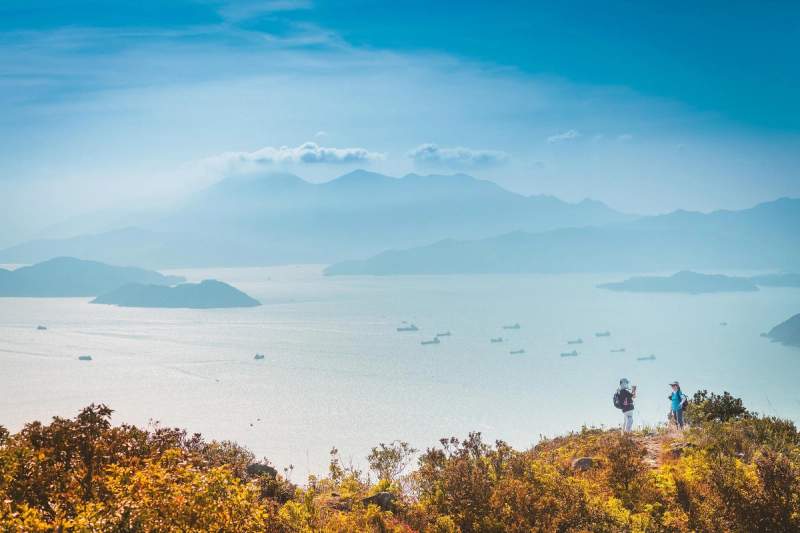 Randonneurs sur une colline surplombant une vue panoramique sur des îles et l'océan, avec des bateaux au loin sous un ciel bleu clair.