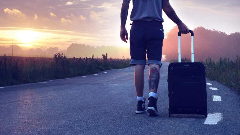 Voyageur marchant sur une route au lever du soleil, tirant une valise, symbolisant l'aventure et la liberté en voyage.