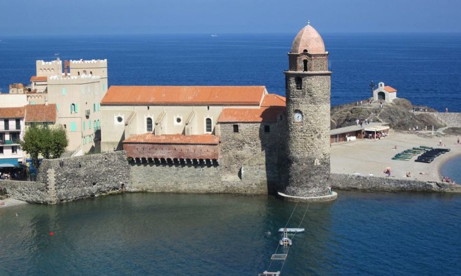 Vue sur le clocher de l’église Notre-Dame-des-Anges à Collioure, en bord de mer Méditerranée, avec plage et promeneurs sous un ciel bleu