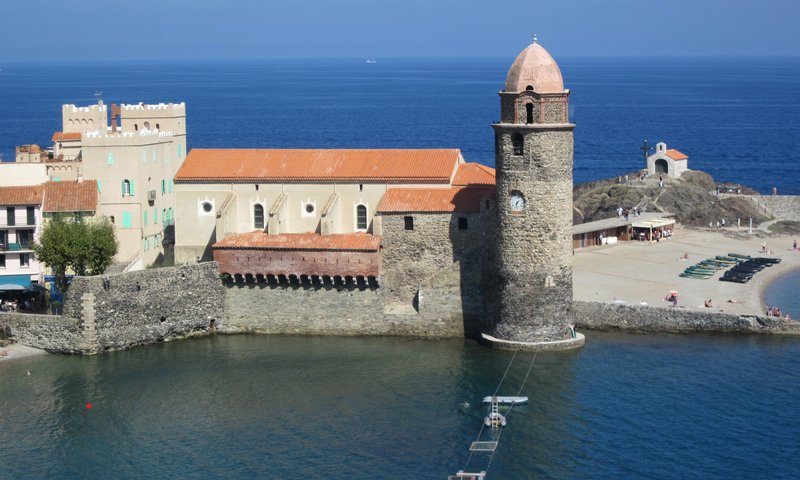 Vue sur le clocher de l’église Notre-Dame-des-Anges à Collioure, en bord de mer Méditerranée, avec plage et promeneurs sous un ciel bleu