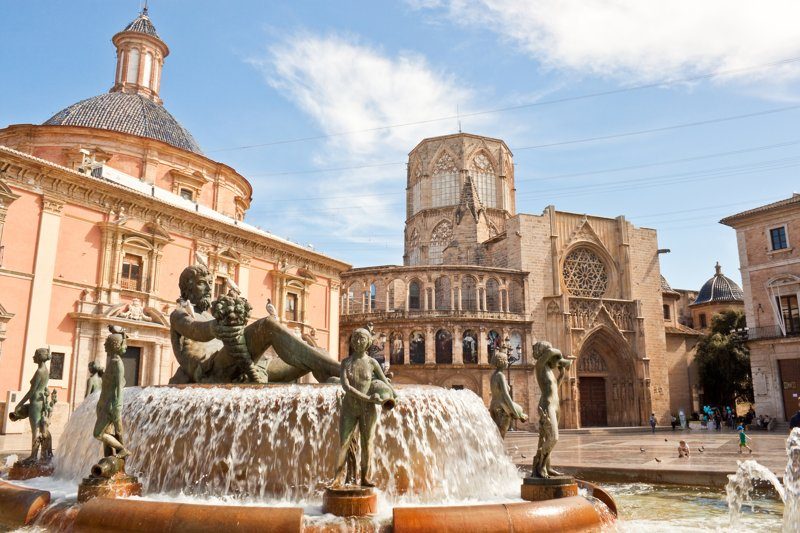 Fontaine de la Plaza de la Virgen dans la vieille ville de Valence, avec la basilique et la cathédrale en arrière-plan, sous un ciel ensoleillé.