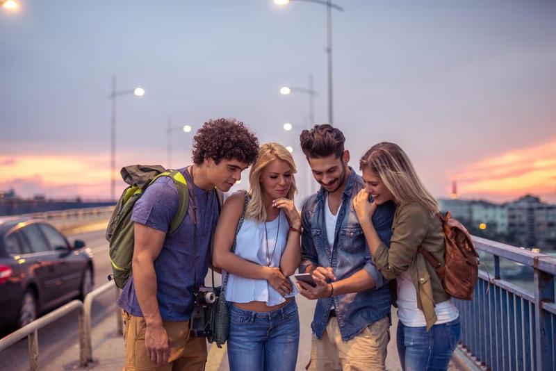 Groupe de quatre amis voyageurs consultant un smartphone sur un pont urbain au coucher du soleil