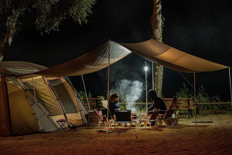 Deux personnes assises sous un abri de camping la nuit, autour d’un feu, à côté d’une grande tente éclairée par une lampe.