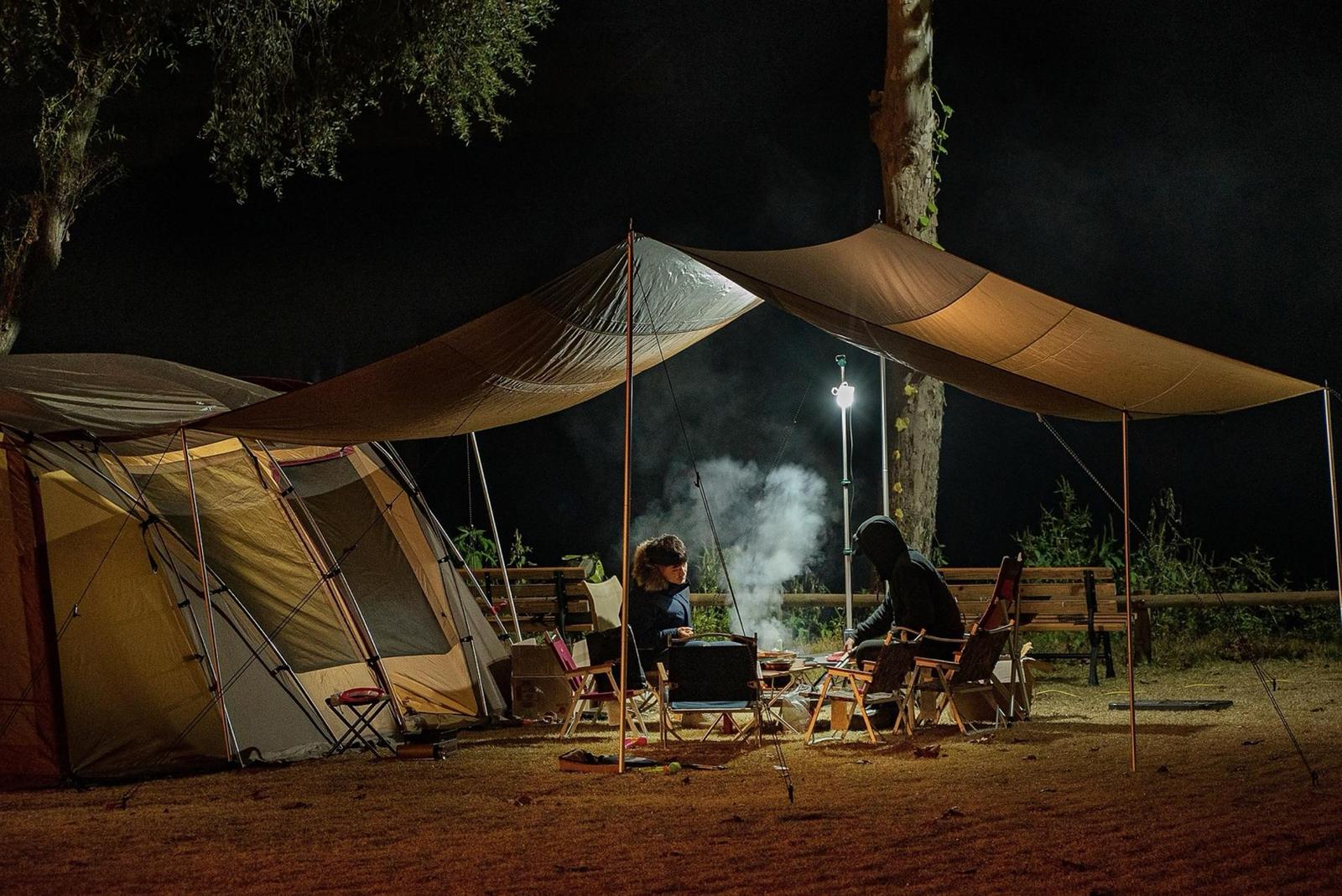 Deux personnes assises sous un abri de camping la nuit, autour d’un feu, à côté d’une grande tente éclairée par une lampe.