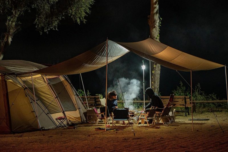 Deux personnes assises sous un abri de camping la nuit, autour d’un feu, à côté d’une grande tente éclairée par une lampe.