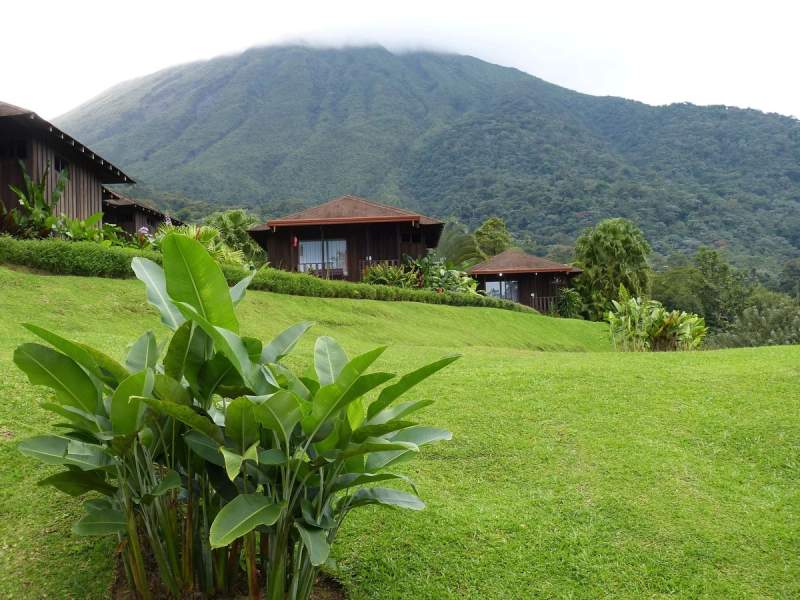 Chalets en bois au pied d'une montagne verdoyante au Costa Rica, avec des plantes tropicales en premier plan et un volcan partiellement couvert de nuages en arrière-plan.