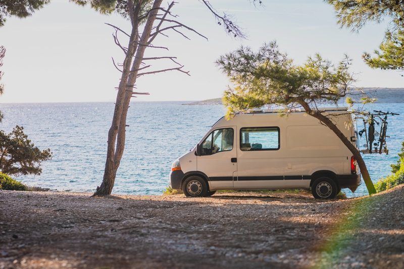 Van aménagé stationné en bord de mer sous des pins, avec porte-vélos à l’arrière, en voyage nomade.