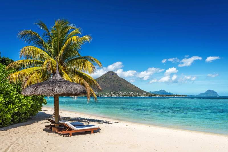 Plage paradisiaque à l'Île Maurice avec son sable blanc, une eau turquoise et un cocotier ombrageant un transat sous un ciel bleu éclatant.