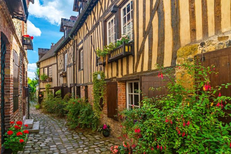 Ruelle pavée bordée de maisons à colombages typiques de Normandie, avec des fleurs aux fenêtres et sur les murs.
