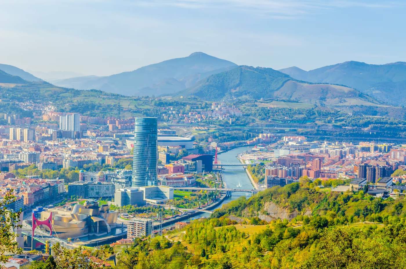 Vue panoramique de la ville de Bilbao, avec le musée Guggenheim et la tour Iberdrola en premier plan, entourés de montagnes verdoyantes.