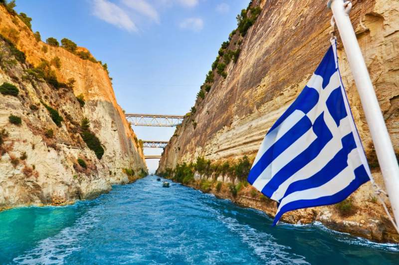 Vue du canal de Corinthe en Grèce, avec une falaise rocheuse et le drapeau grec flottant au premier plan.