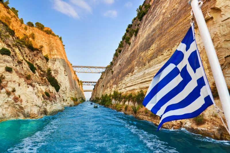 Vue du canal de Corinthe en Grèce, avec une falaise rocheuse et le drapeau grec flottant au premier plan.